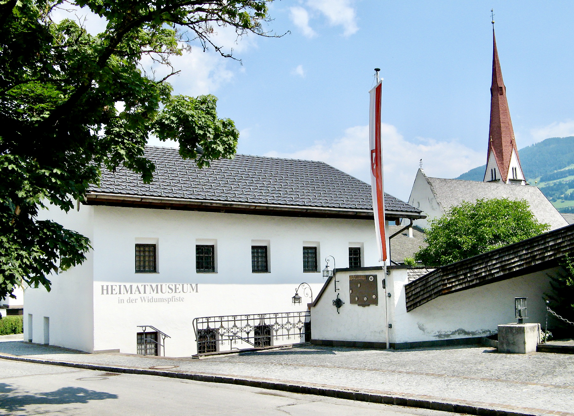 Foto des Heimatmuseums in der Widumspfiste mit Kirchenturm und Bannerflagge vor blauem Himmel.