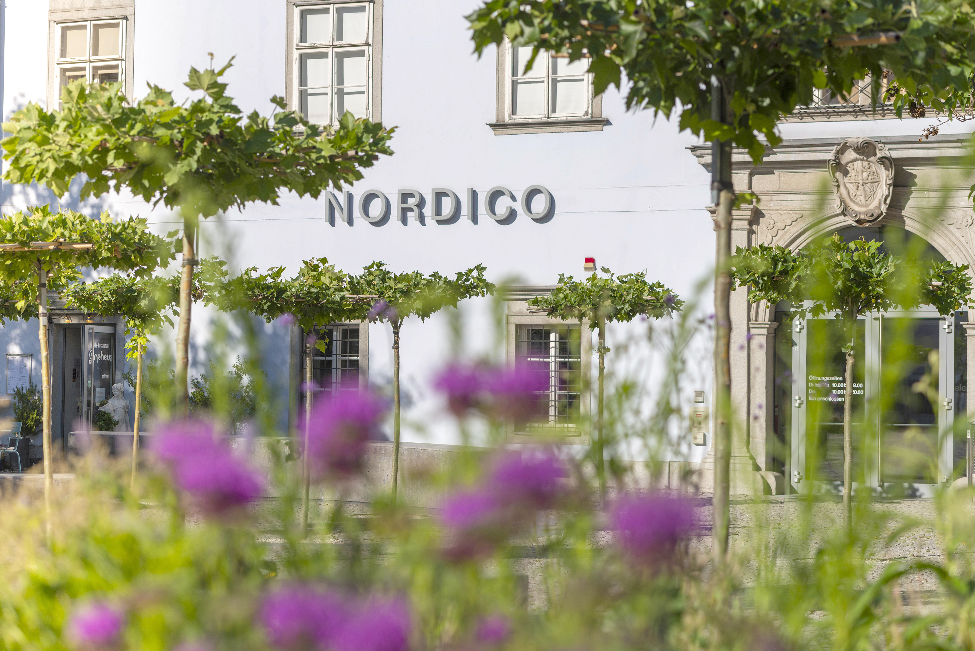 Blick auf die Fassade des Nordico Stadtmuseums durch den begrünten und blühenden Vorplatz.