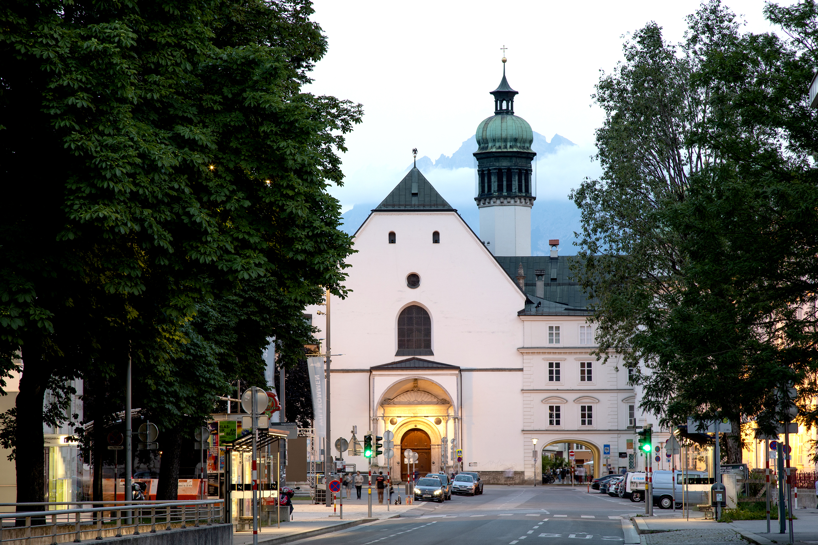Blick auf die Hofkirche Innsbruck entlang der dorthin führenden Straße. Auf dem Foto ist die Kirche von den auf den beiden Straßenseiten stehenden Bäumen eingerahmt.
