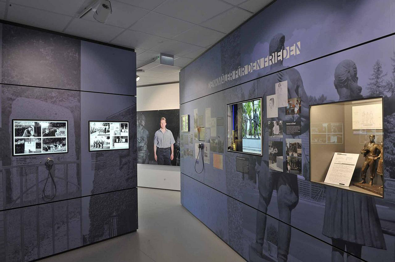 Foto eines Ausstellungsraums im "Erlauf erinnert" Museum. Zu sehen sind Dokumente und Bronzestatuen in einem Schaukasten, und diverses Bildmaterial.