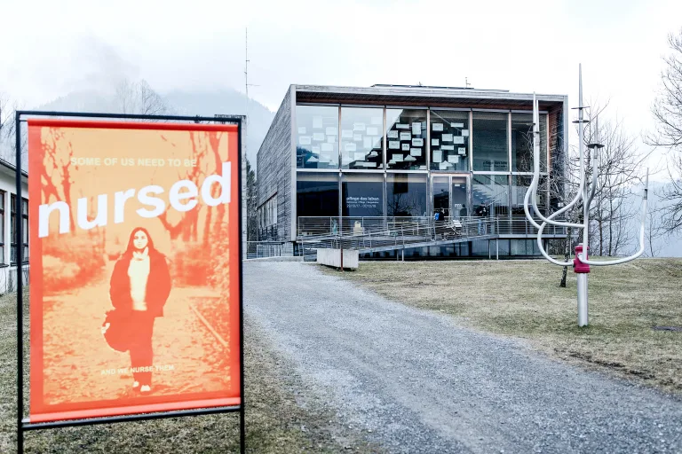 Foto des quaderförmigen Frauenmuseums Hittisau bei bewölktem Himmel. Neben dem Schotterweg zum Museum hängt ein Plakat, das Frauen in der Pflege thematisiert.