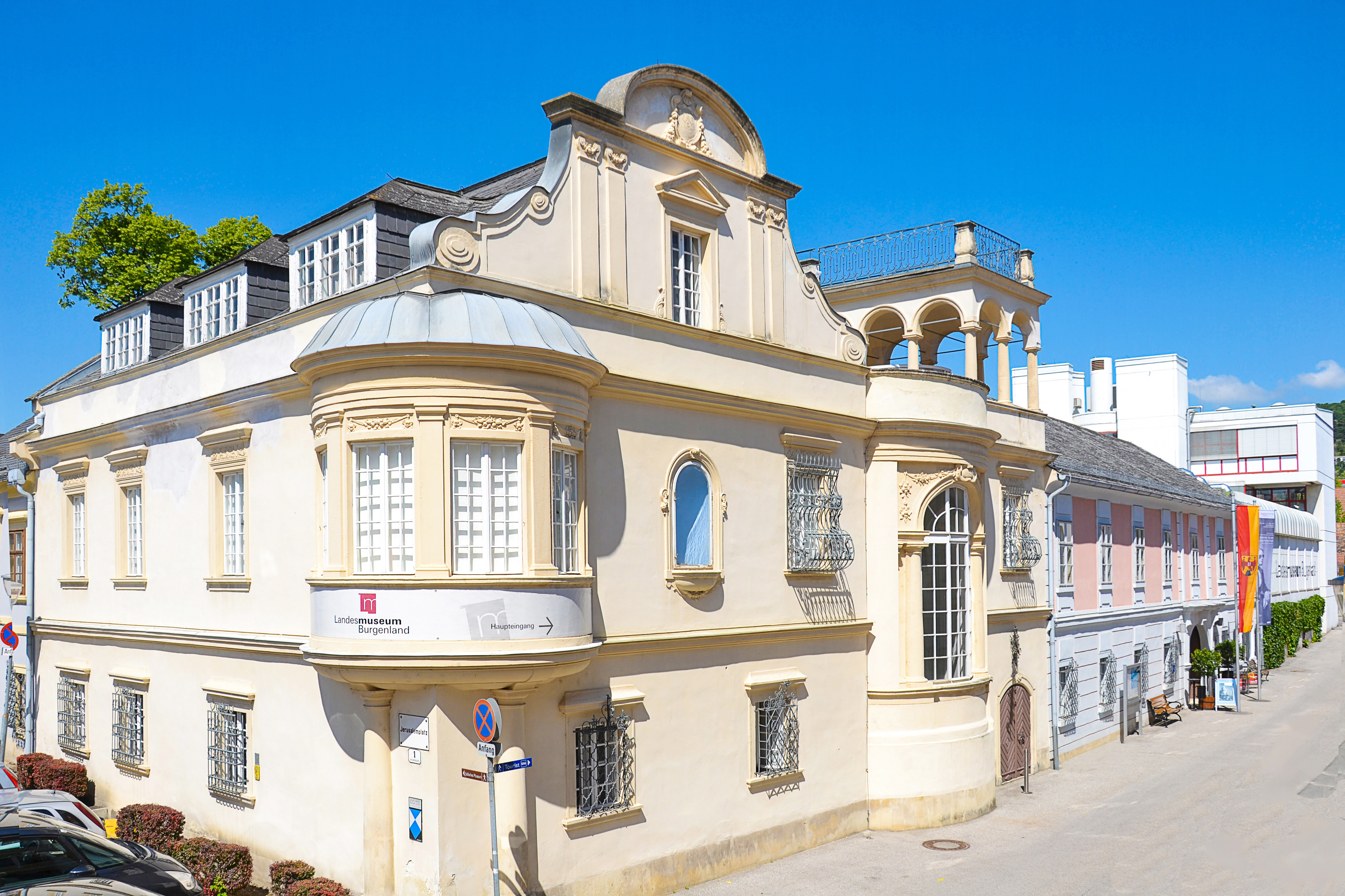 Blick auf das beigefarbene Landesmuseum Burgenland in Eisenstadt bei strahlend blauem Himmel.