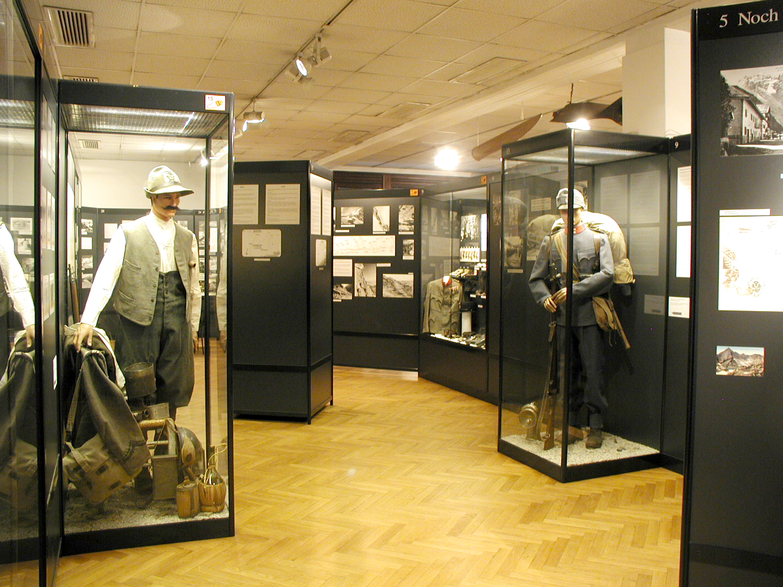 Foto eines Ausstellungsraums im Museum 1915-1918. Gezeigt werden typische Bekleidung und Ausrüstung von Zivilisten und Soldaten an in Vitrinen aufgestellten Schaufensterpuppen.