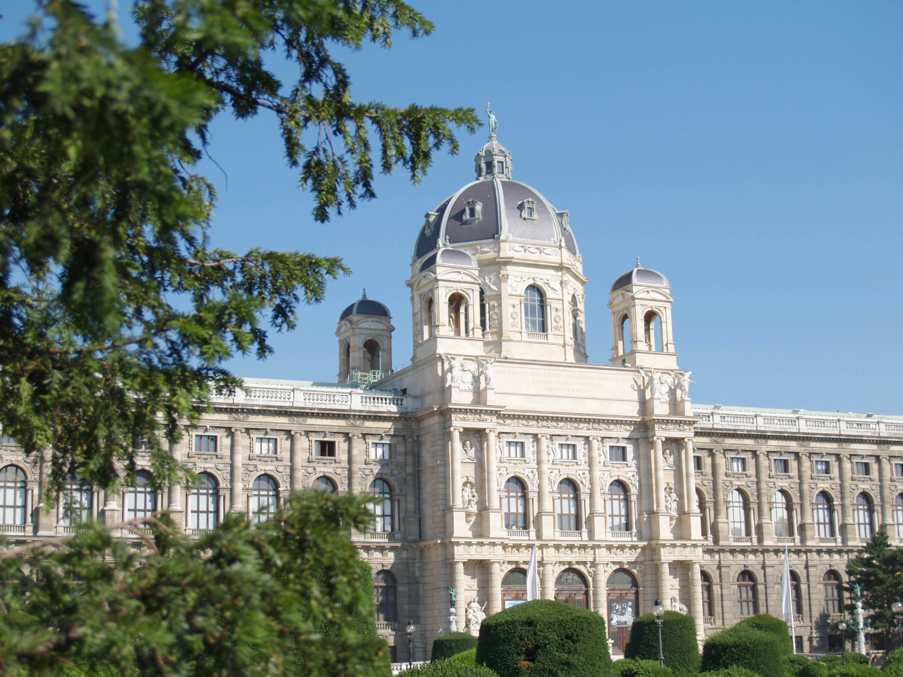 Blick durch den Vorgarten auf das naturhistorische Museum Wien bei hellblauem Himmel.