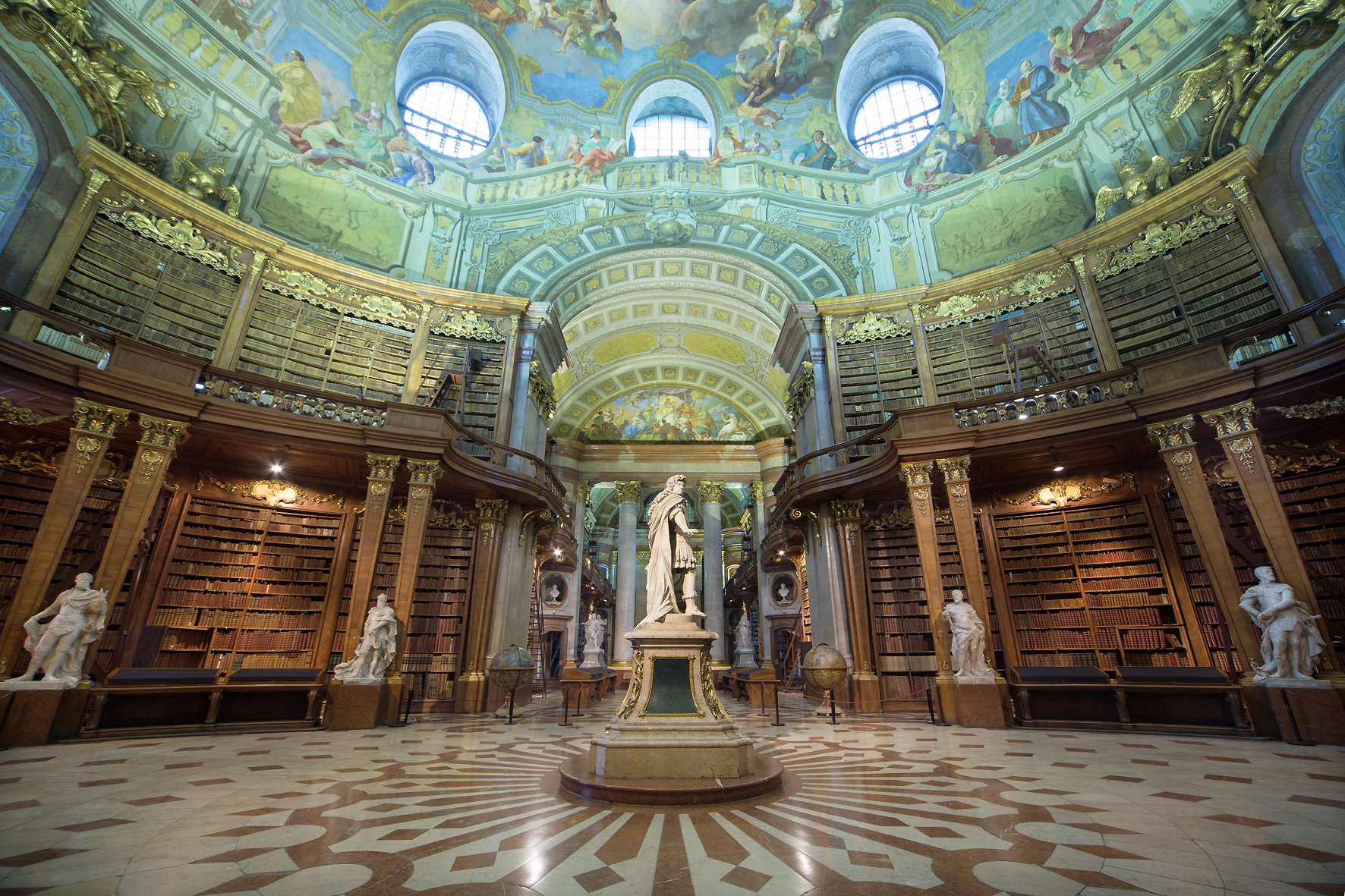 Foto des Prunksaals der Österreichischen Nationalbibliothek. Zu sehen sind mehrere Marmorfiguren, Bodenverzierungen, Goldornamente an den Wänden und Bücherregalen, Deckengemälde und zwei Globen. An den Wänden stehen große mit ledergebundenen Büchern gefüllte Bücherregale.