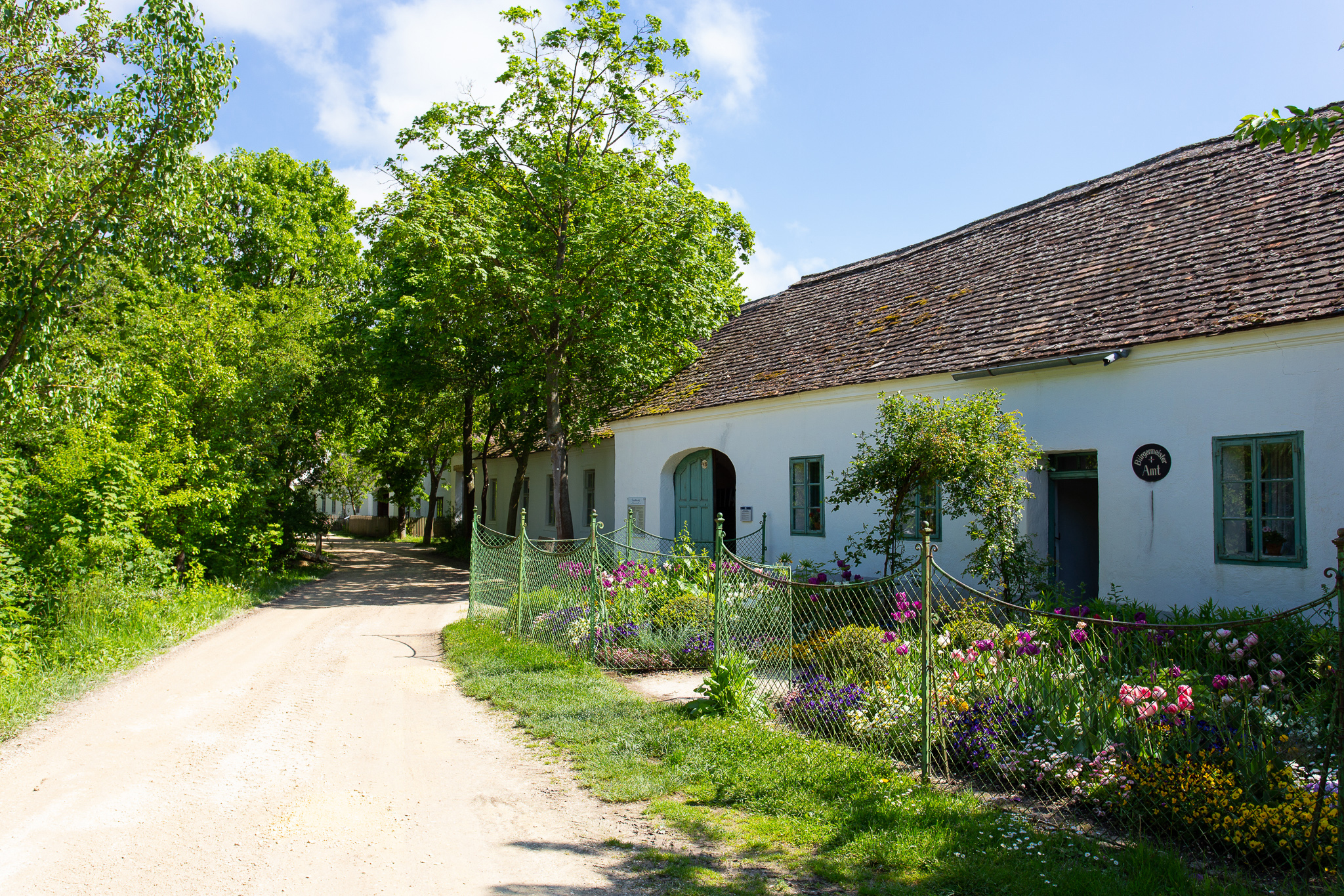 Von Bäumen eingerahmter Schotterweg und blühender Blumengarten vor einem einstöckigen, langgezogenen Haus.