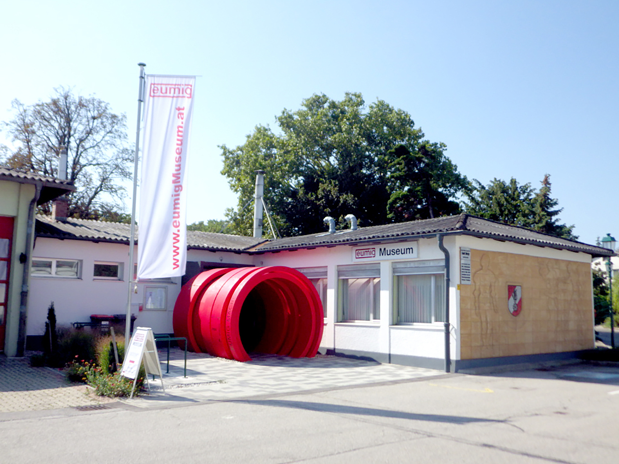 Foto des eumigMuseums mit Fahne und markantem rotem tunnelartigen Objekt vor dem Gebäude.