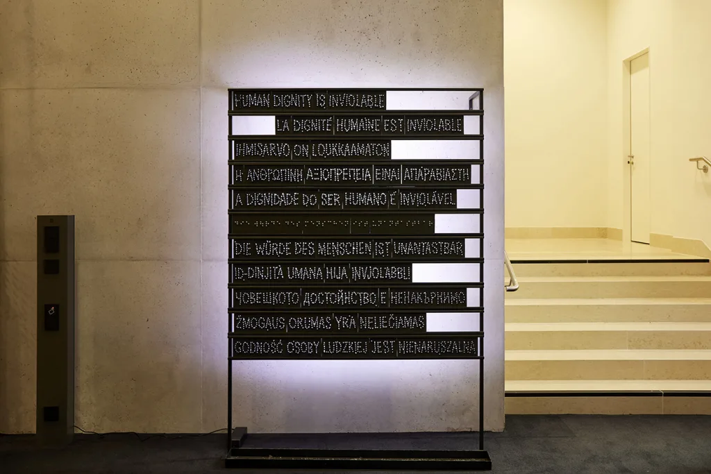 Foto einer Installation im Österreichischen Parlament, auf der der erste Satz aus Artikle 1 der Charta der Grundrechte der Europäischen Union in verschiedenen Sprachen und auch in Brailleschrift abgebildet wird.