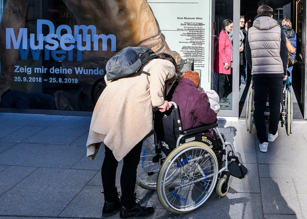 Zu sehen sind mehrere Menschen im Eingangsbereich des Dom Museums Wien. Zwei Personen befinden sich im Rollstuhl und werden von Assistenzpersonen geschoben.
