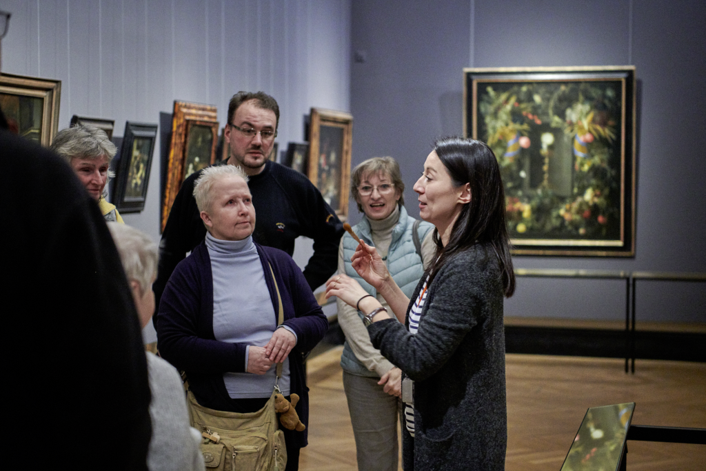 Foto einer Gruppe von Menschen bei einer Führung in einfacher Sprache im Kunsthistorischen Museum Wien. Im Hintergrund ist eine Reihe von eingerahmten Gemäden zu sehen.