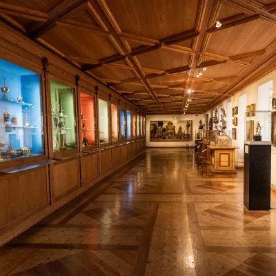 Zu sehen sind diverse Ausstellungsobjekte entlang eines Ausstellungsgangs im Ambraser Unterschloss, Innsbruck. Die linke Vitrinenwand, sowie der Parkettboden und die Decke sind aus Holz, was dem Raum ein besonderes Ambiente verleiht.