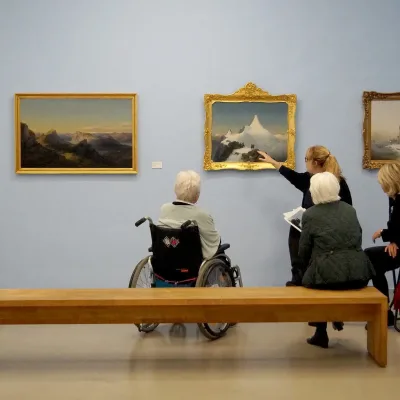 Foto einer Führung im Museum Moderner Kunst Kärnten. Zu sehen sind fünf Personen, die zum Teil im Rollstuhl oder am Rollator sitzend den Erläuterungen zu einem Gemälde eines schneebedeckten Berges lauschen.