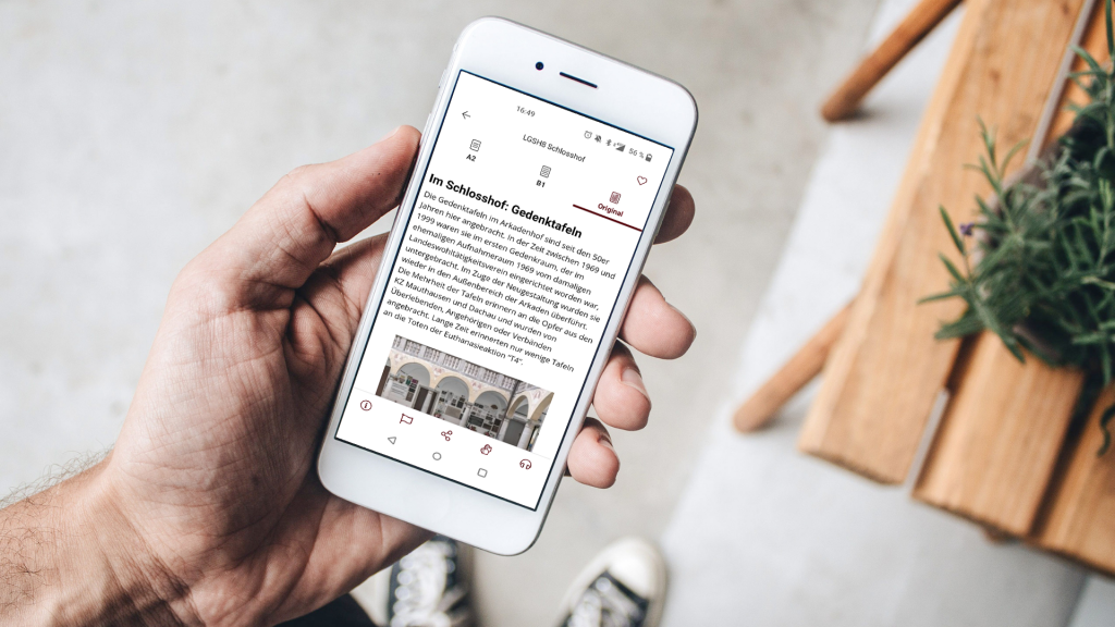 Foto eines von einer Männerhand gehaltenen Smartphones. Auf dem Bildschirm ist ein Ausschnitt zu den Gedenktafeln im Arkadenhof zu sehen, wie er in der capito App dargestellt wird.