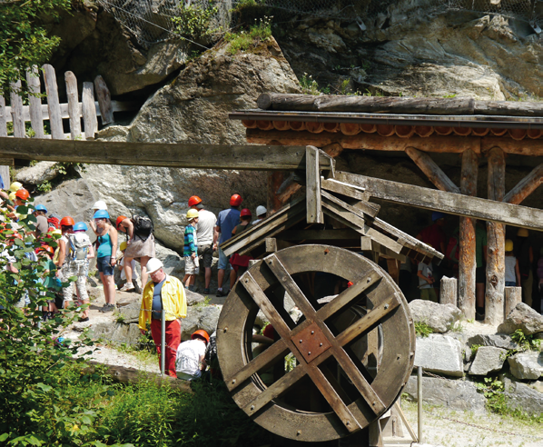 Vor einer Felswand steht ein hölzernes Wasserrad. Eine Schulgruppe mit Helmen unterschiedlicher Farbe hört einem Vortrag zu.