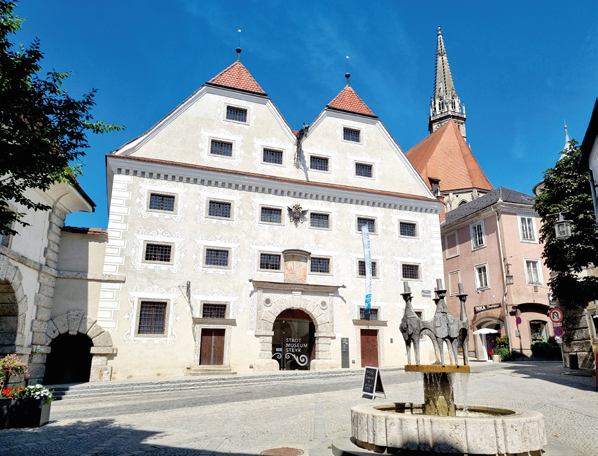 Das Foto zeigt den Brunnen vor dem Stadtmuseum Steyr sowie das Haus, in dem sich das Museum befindet, an einem schönen Tag.