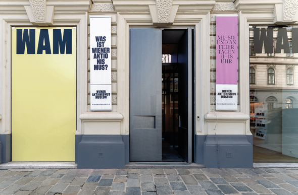 Foto des Eingangs zum Wiener Aktionismus Museum. Auf einem gelb gefärbten, bodenhohen Fenster links neben dem Eingang steht in großen schwarzen Buchstaben "WAM".