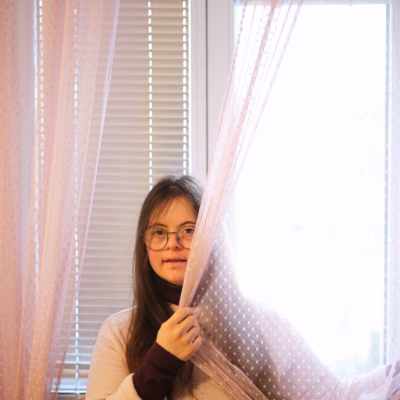 Eine junge Frau mit Brille blickt vor einem Fenster durch einen rosa Vorhang
