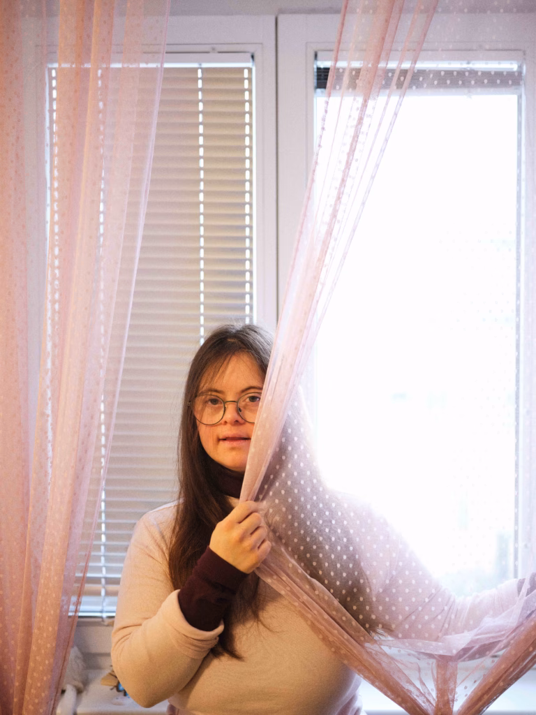 Eine junge Frau mit Brille blickt vor einem Fenster durch einen rosa Vorhang