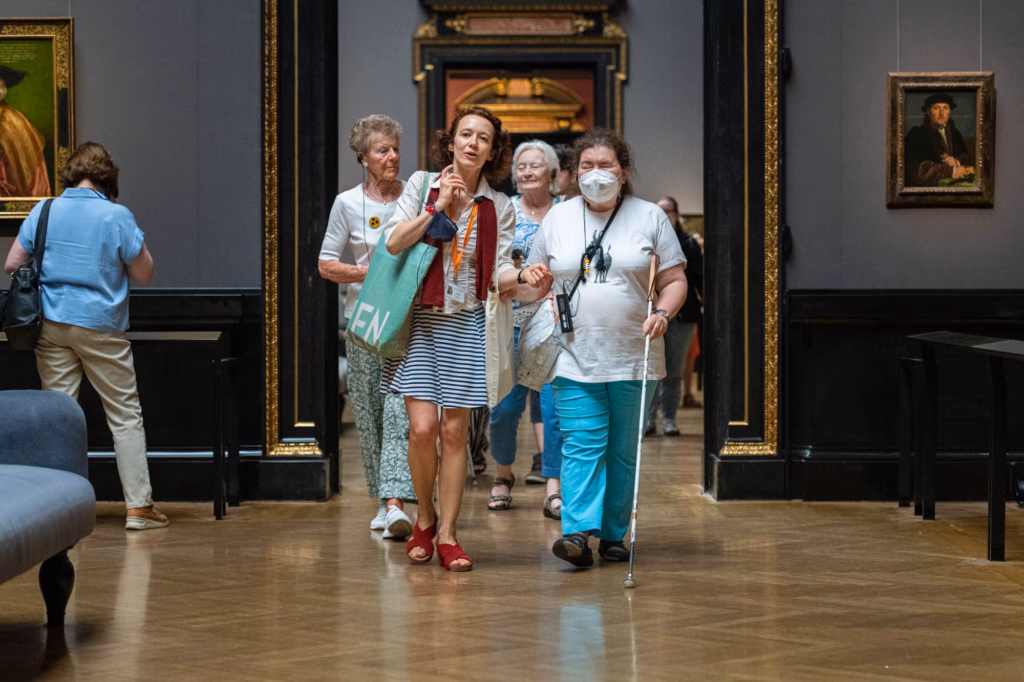 Eine Begleiterin führt eine blinde Museumsbesucherin mit einem Langstock durch den Ausstellungsraum