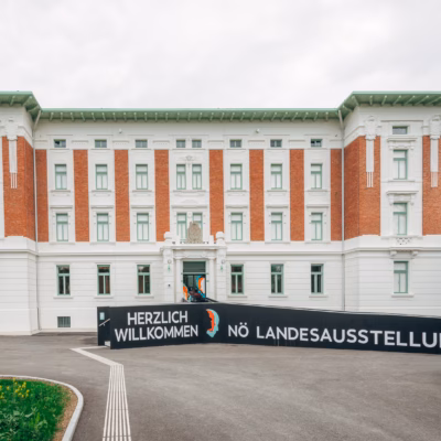 Vorderansicht eines großen historischen Gebäudes mit weißer Fassade und roten Ziegelakzenten vor einem bewölkten Himmel