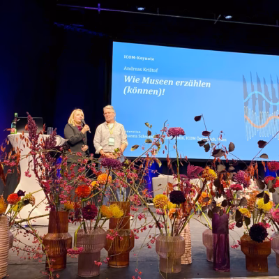 Zwei Personen stehen auf einer Bühne vor einer Leinwand mit der Aufschrift „Wie Museen erzählen (können)!“, davor stehen farbenfrohe Blumenarrangements in Vasen